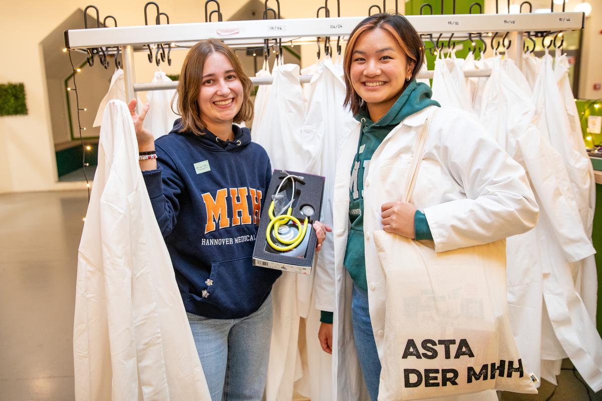 Copyright: Karin Kaiser / MHH zwei Frauen stehen in einer Garderobe, an der viele weiße Kittel hängen und präsentieren die Ersti-Beutel und deren Inhalt.