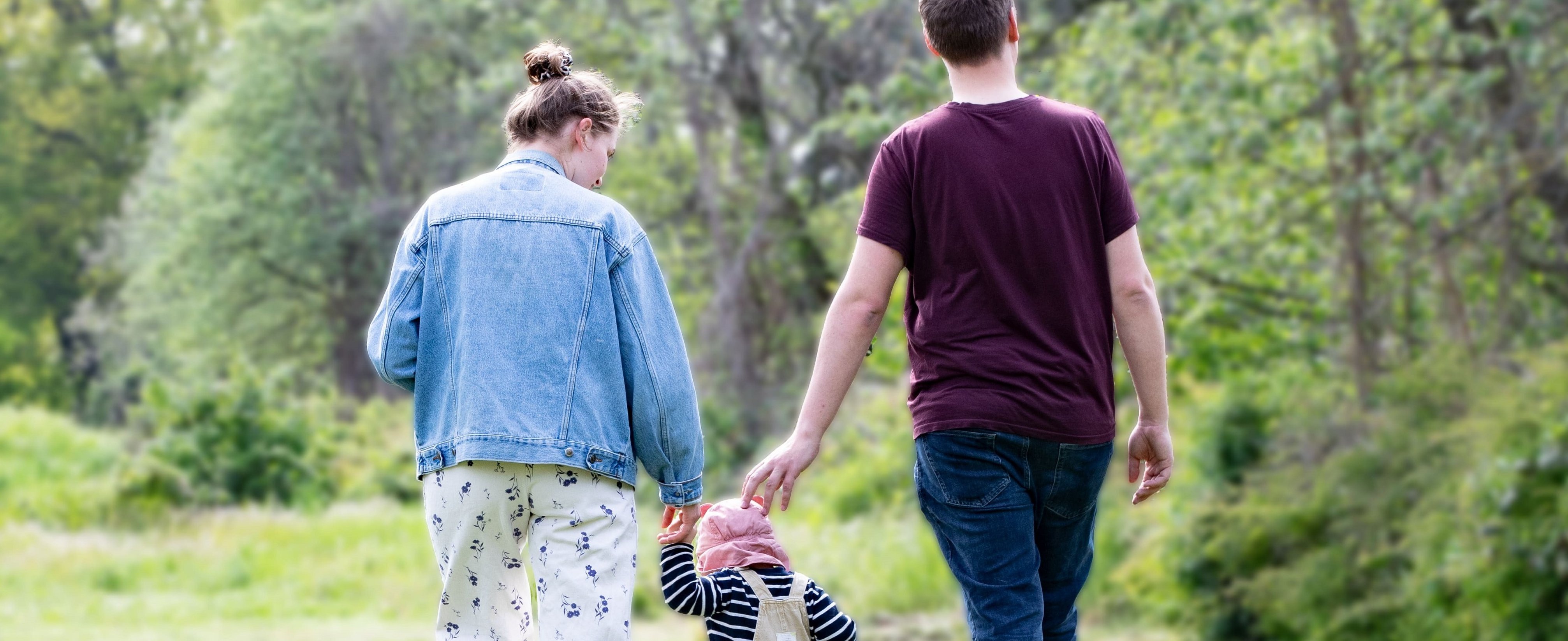 A man and a woman are walking side by side across a meadow, holding a small child in their hands.