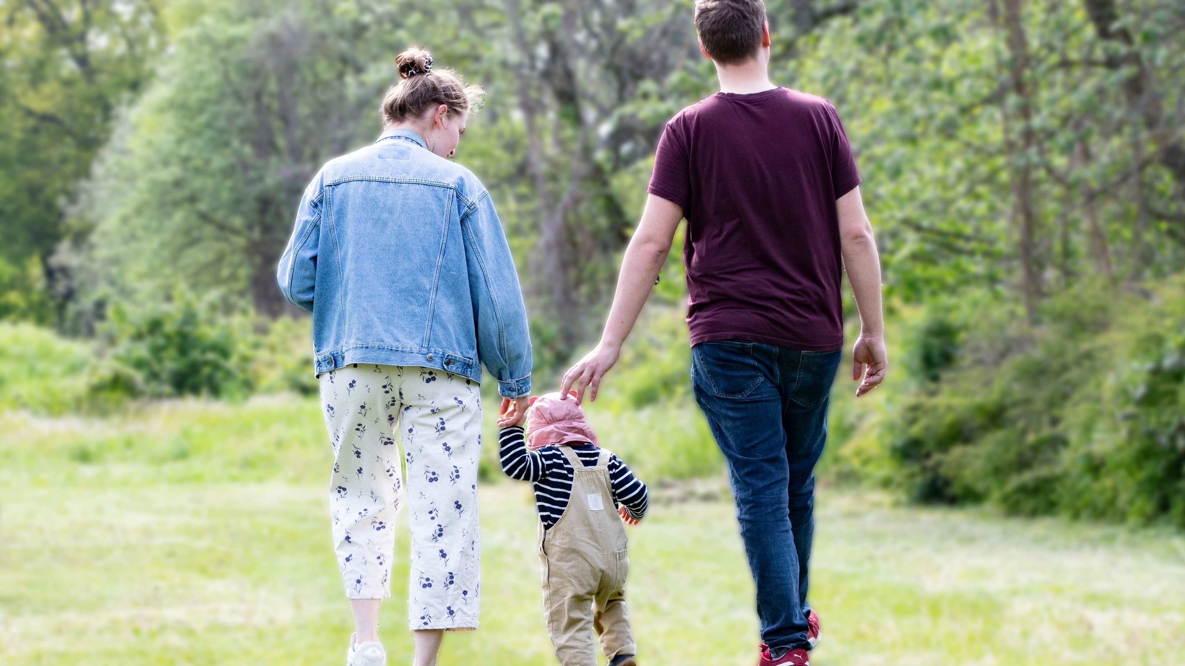 A woman and a man are walking on a green meadow, with a small child running in their midst. They can all be seen from behind.