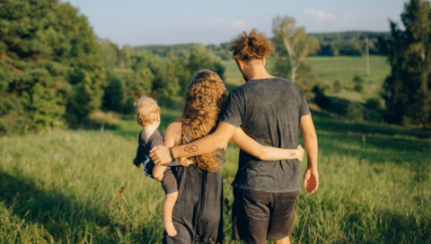 A woman and a man are holding each other, their backs to the camera. The woman is holding an infant. Both woman and man have the organ donation tattoo.