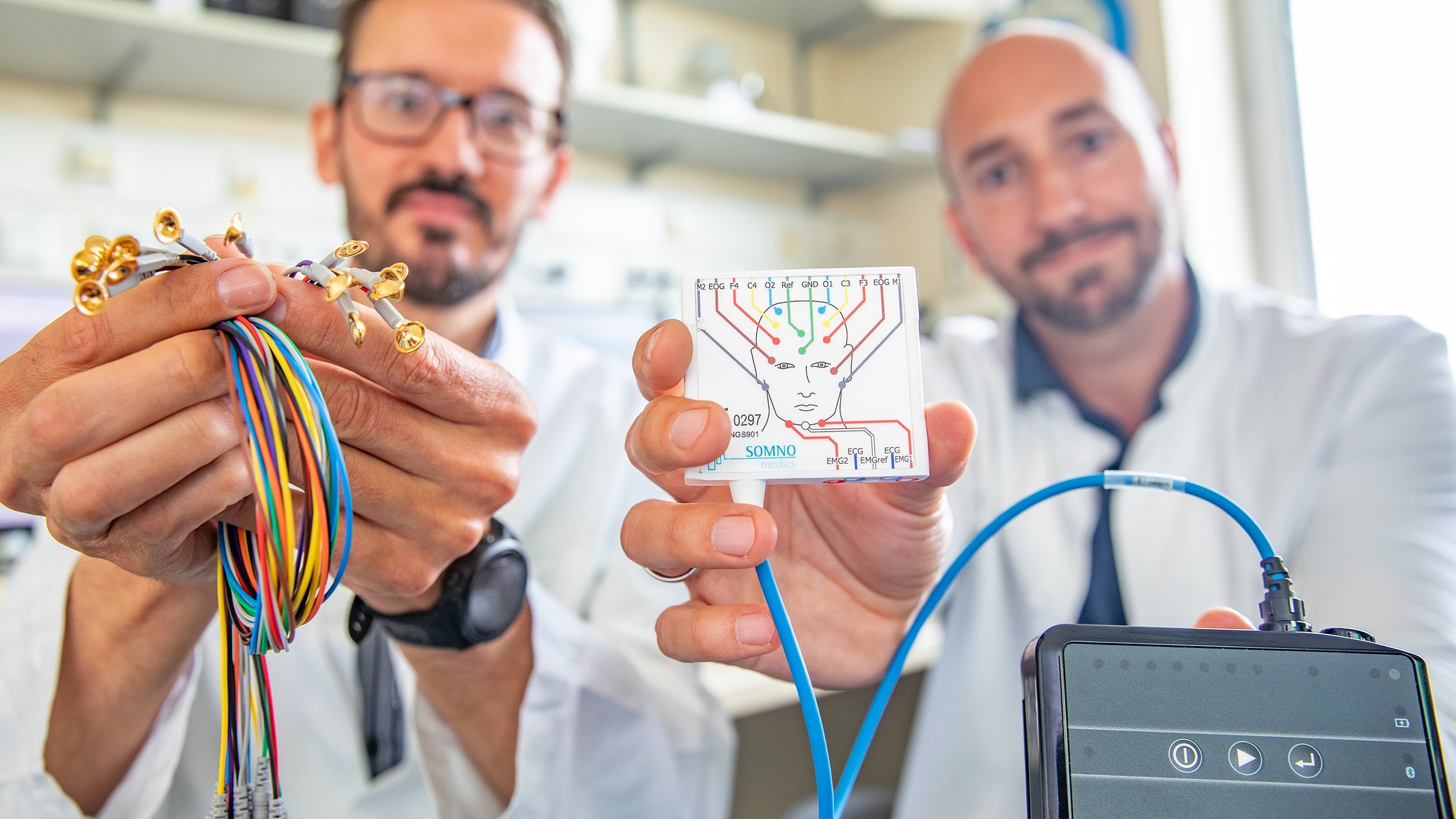 Dr. Klietz, der rechts sitzt, hält ein eckiges schwarzes Gerät in die Kamera, aus dem ein blaues Kabel zu eienr Platte führt, auf dem ein menschlicher Kopf abgebildet ist. Dr. Höllerhage hält ein Bündel von bunten Kabeln in der Hand.