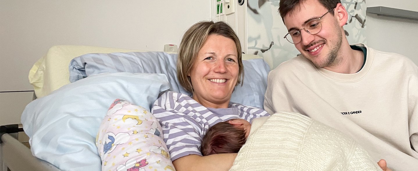 Mutter Ann-Merlin und Vater Paul liegen mit ihrem Neugeborenen in einem Krankenhausbett. 