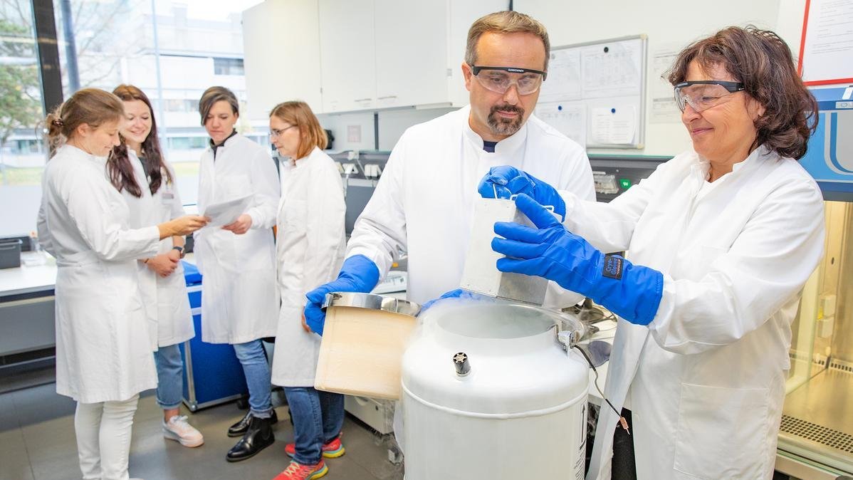 Professor Dr. Eiz-Vesper and Professor Dr. Thomas Skripuletz take a package of frozen T-cells out of a special cryo container. Four employees are standing in the background.