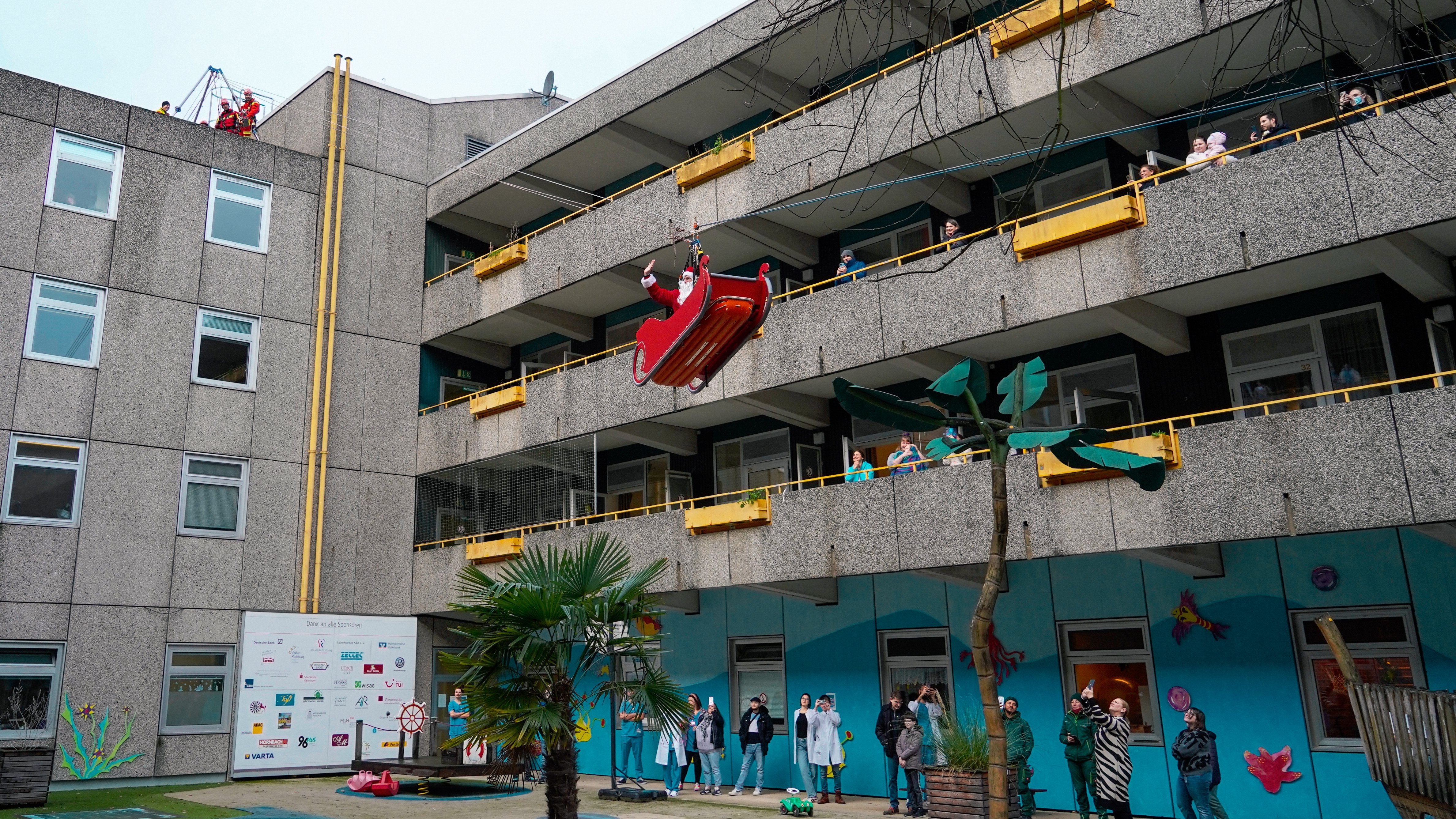 Ein Höhenretter in Nikolauskostüm wird in einem Schlitten an der Fassade der MHH-Kinderklinik abgeseilt. Unten und auf den Balkonen schauen ihm Menschen zu. 