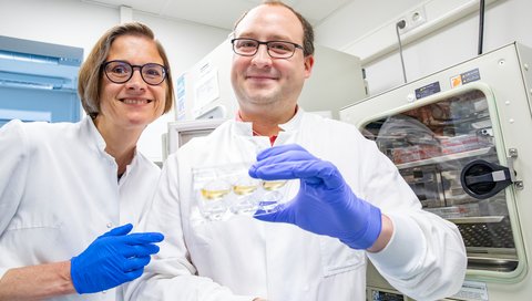 Eine Frau und ein Mann in weißem Kittel stehen in einem Labor und zeigen Zellkulturen.