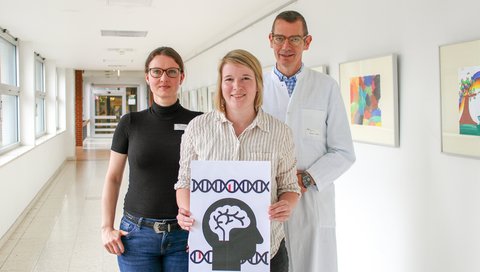 Three researchers in the hallway to the children's clinic. In their hands they hold a simplified illustration of their research, which shows mutated genes and a brain.