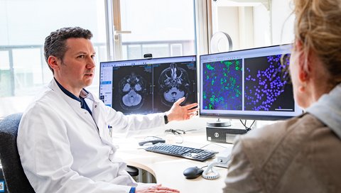 A doctor in a white coat sits in front of two screens with MRI images of the cerebellum and fluorescence microscope images of cell cultures.