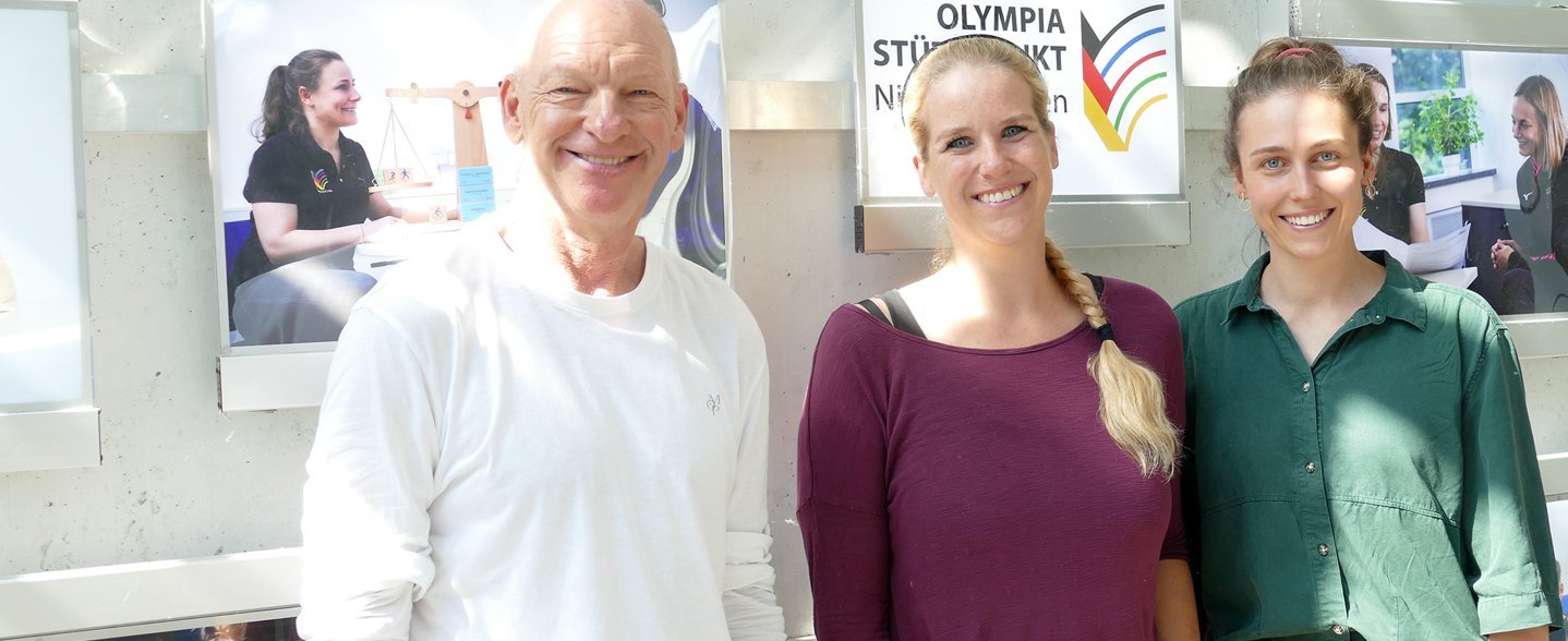 Ein Mann und zwei Frauen stehen vor einer Bilderwand. 