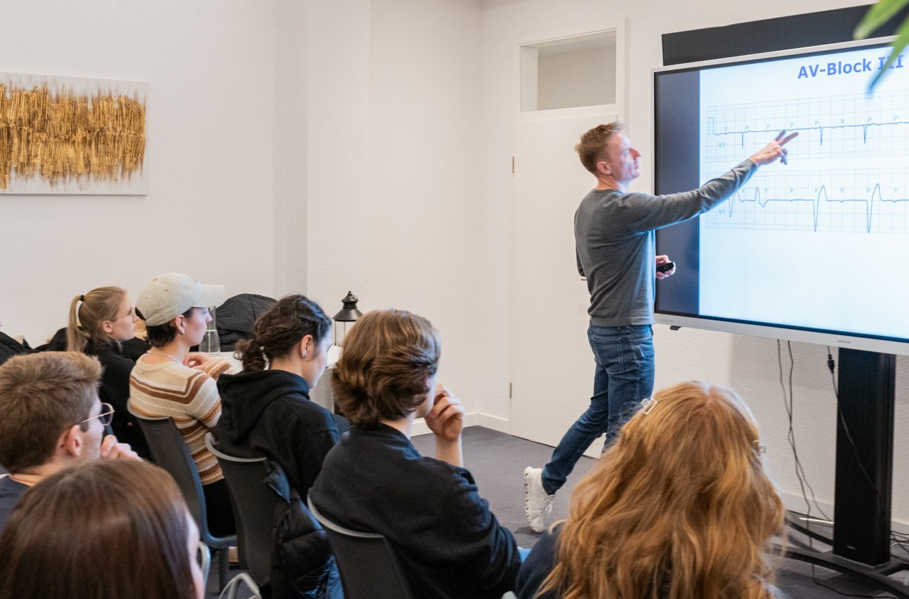 Der Referent zeigt auf ein Whiteboard, Studierende sitzen in Stuhlreihen davor und blicken nach vorne auf das Board.