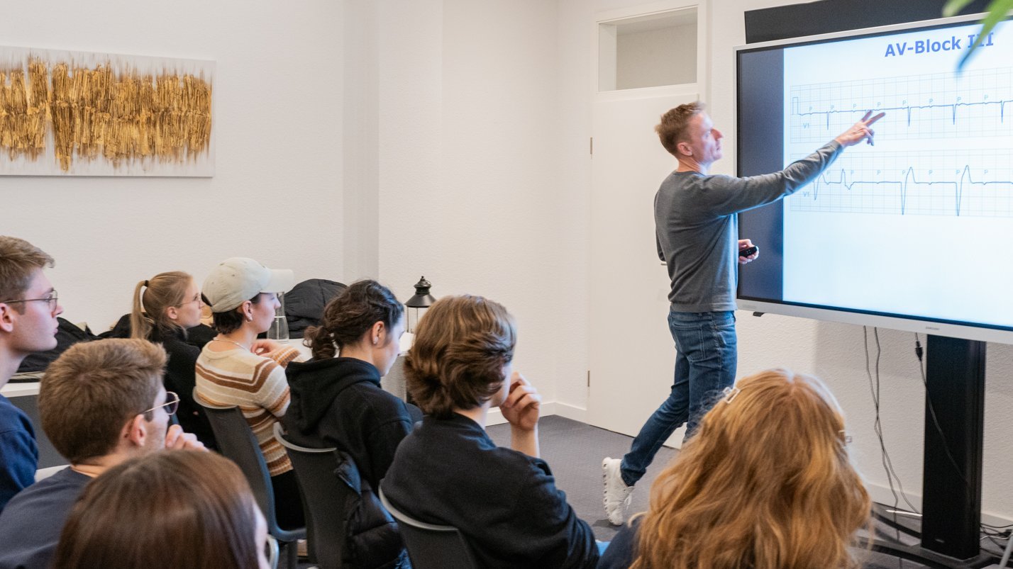 Der Referent zeigt auf ein Whiteboard, Studierende sitzen in Stuhlreihen davor und blicken nach vorne auf das Board.