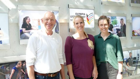 Ein Mann und zwei Frauen stehen vor einer Bilderwand mit dem Schriftzug Olympia Stützpunkt Niedersachsen. . 