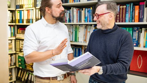 Zwei Männer stehen mit einem Buch in der Hand vor einem Bücherregal.