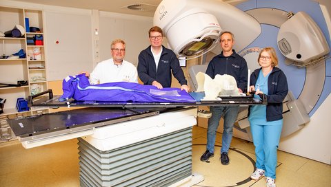 A four-person radiotherapy team stands at a linear accelerator with a rotating table top.