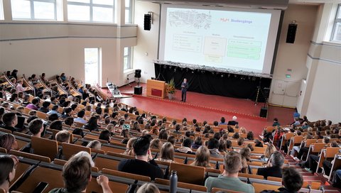 View from above into lecture hall F at MHH. Many young people are sitting in the rows, one person is standing at the front at the desk.