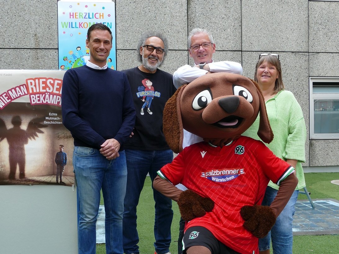 96 Sportdirektor Marcus Mann, David Kadell, Prof. Beerbaum und Annette Wiens stehen nebeneinander im Innenhof der Kinderklinik. Vor ihnen steht das 96-Maskottchen. 