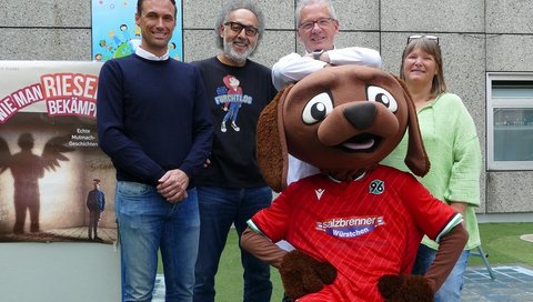 96 Sportdirektor Marcus Mann, David Kadell, Prof. Beerbaum und Annette Wiens stehen nebeneinander im Innenhof der Kinderklinik. Vor ihnen steht das 96-Maskottchen. 