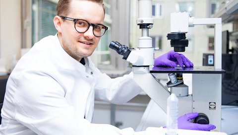 Der Gastroenterologe Dr. Bernd Heinrich sitzt neben einem Mikroskop und hält einen Probenbehälter mit Lebergewebe in der Hand.