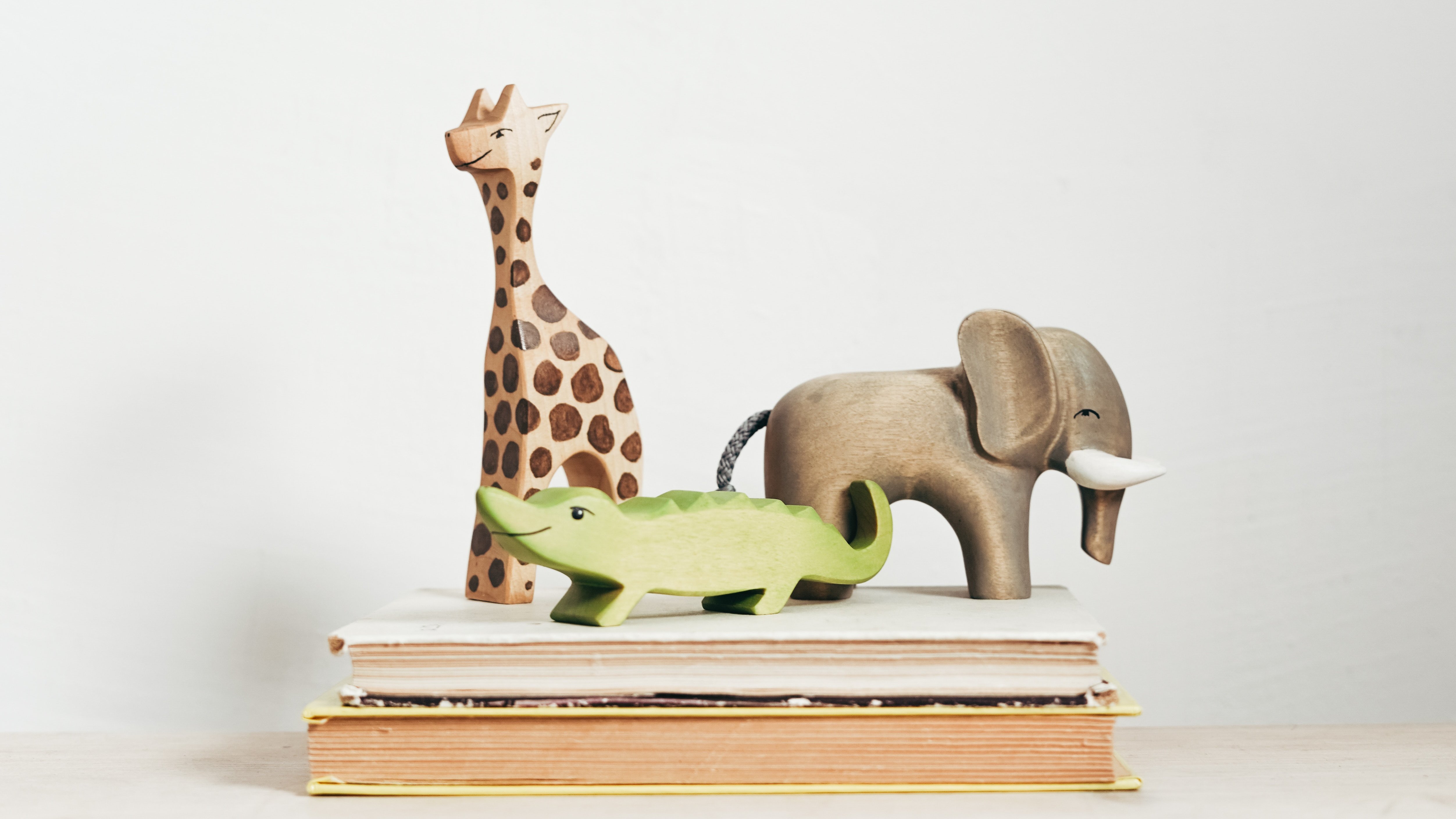 This picture shows three friendly looking wooden toys (a giraffe, an elephant and a crocodile) on two stacked books.