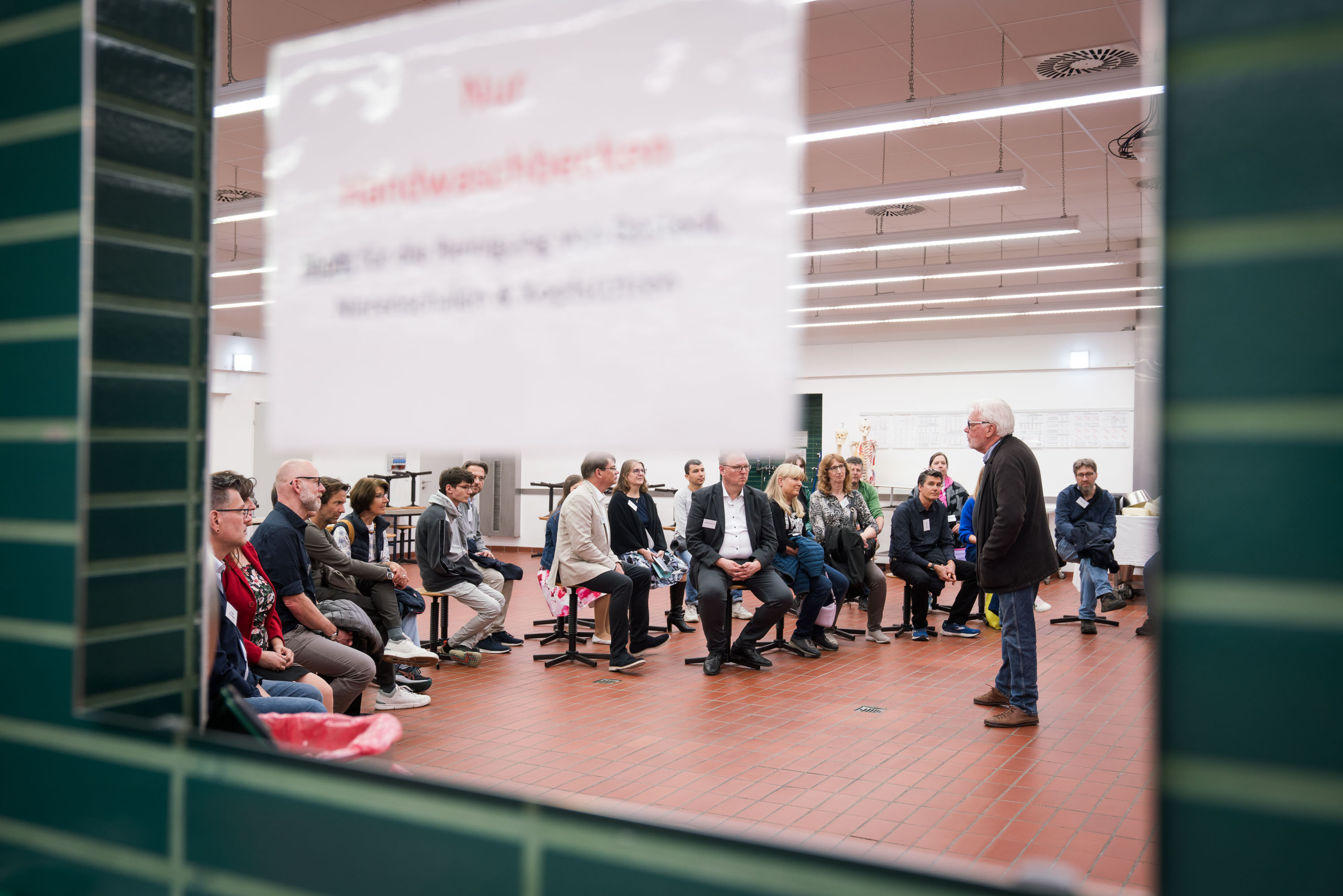 Durch eine Glasscheibe sieht man die Gruppe auf Hockern sitzen, Professor Bartels erzählt etwas.