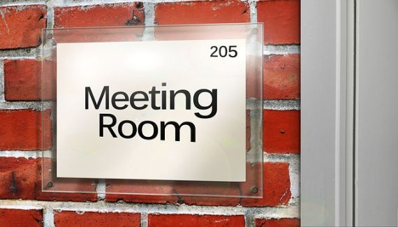 Eine rote Backsteinmauer, darauf ein transparentes Türschild, auf dem Meeting Room steht. Rechts im Bild sieht man noch einen Teil des weißen Türrahmens.