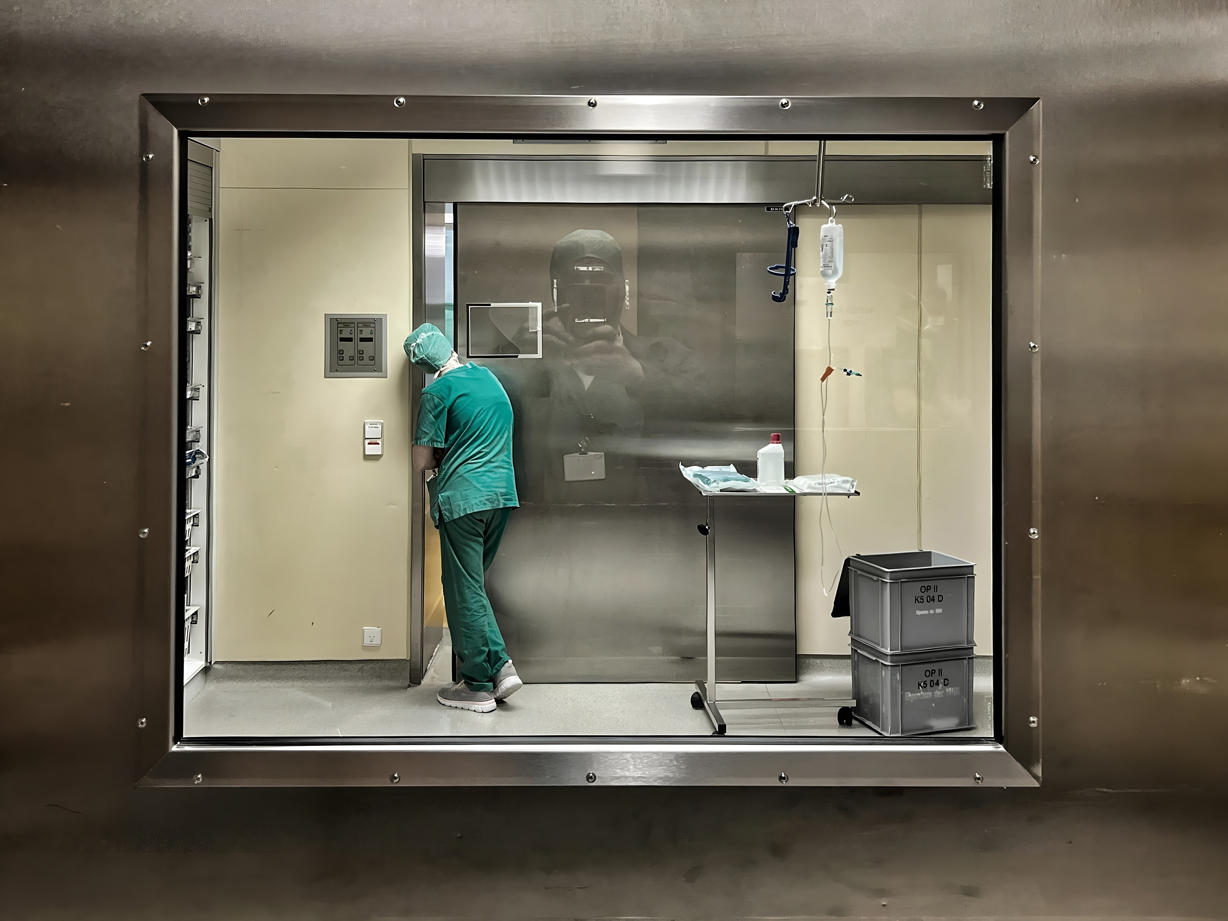 A person in a green coat stands at a metal sliding door to an operating theater and looks in through a small gap.