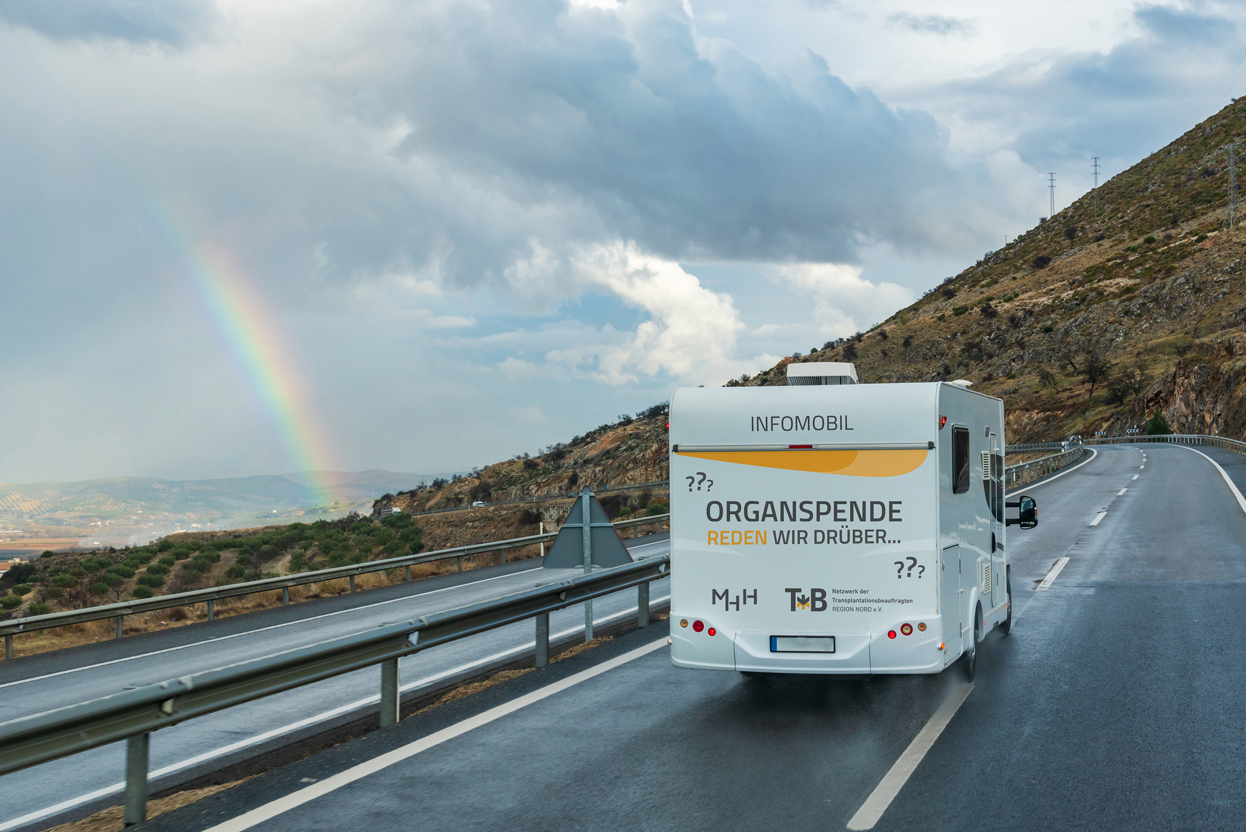 An information camper with the inscription "Organ donation: Let's talk about it" drives along a rain-soaked road; a m horizon reveals a rainbow. 