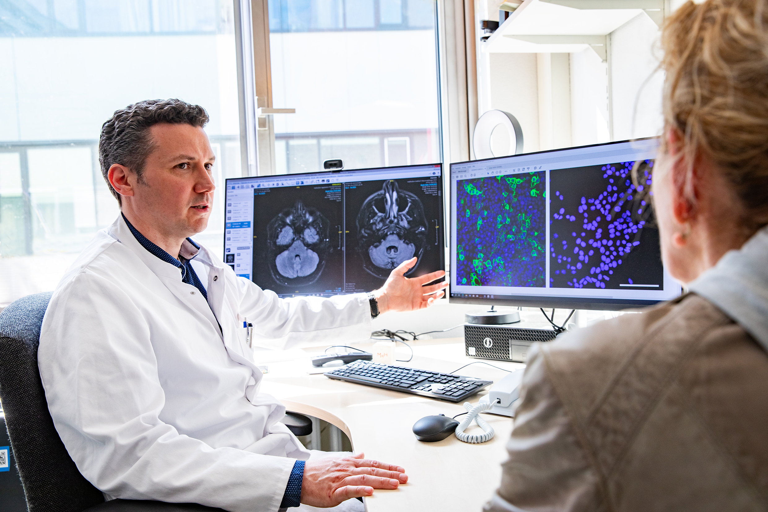 Ein Arzt im weißen Kittel sitzt vor zwei Bildschirmen mit MRT-Aufnahmen des Kleinhirns und fluoreszenzmikroskopischen Bildern von Zellkulturen.