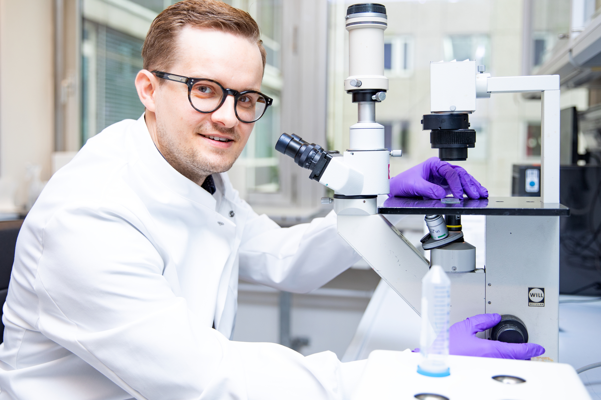 Der Gastroenterologe Dr. Bernd Heinrich sitzt neben einem Mikroskop und hält einen Probenbehälter mit Lebergewebe in der Hand.