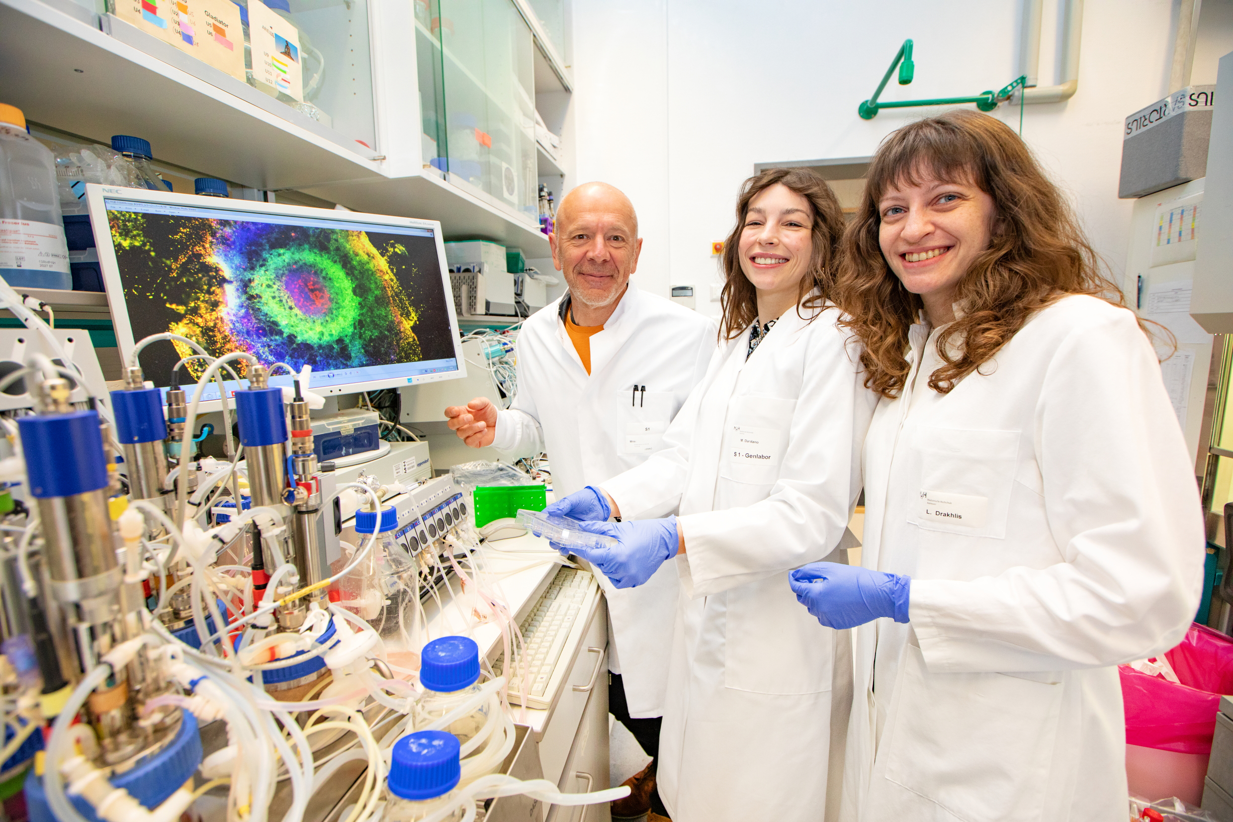 A man and two women stand in front of bioreactors in the laboratory and show a cell culture dish for growing stem cells. 