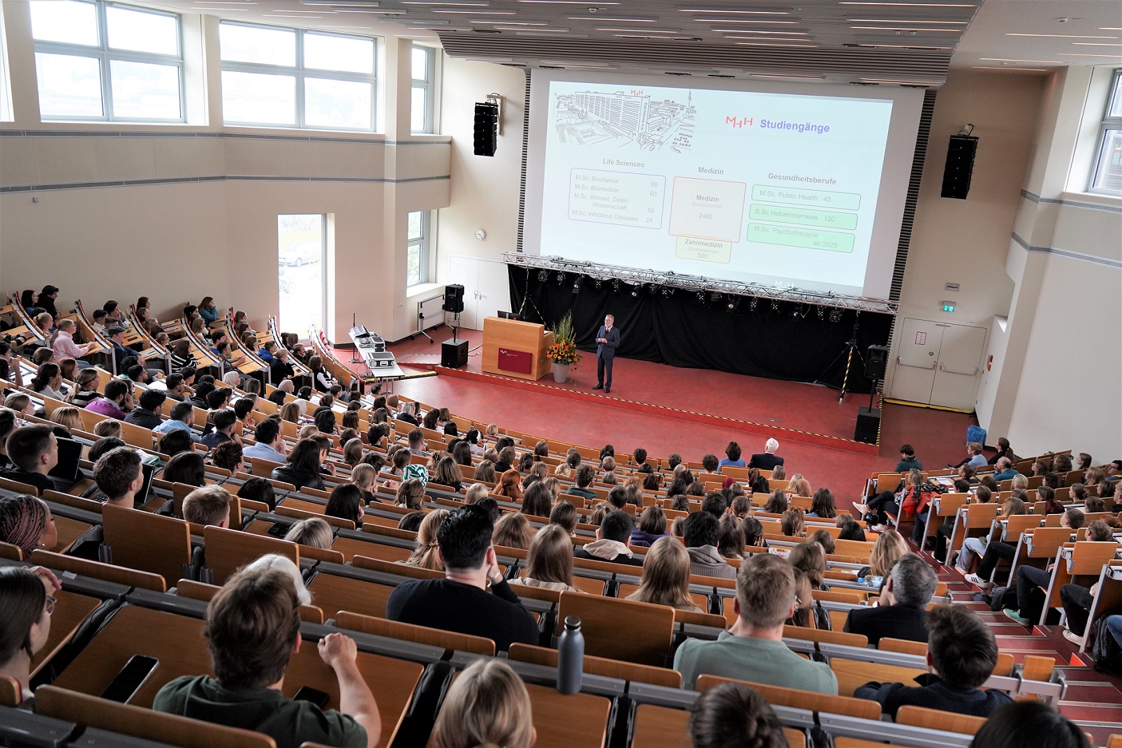 View from above into lecture hall F at MHH. Many young people are sitting in the rows, one person is standing at the front at the desk.