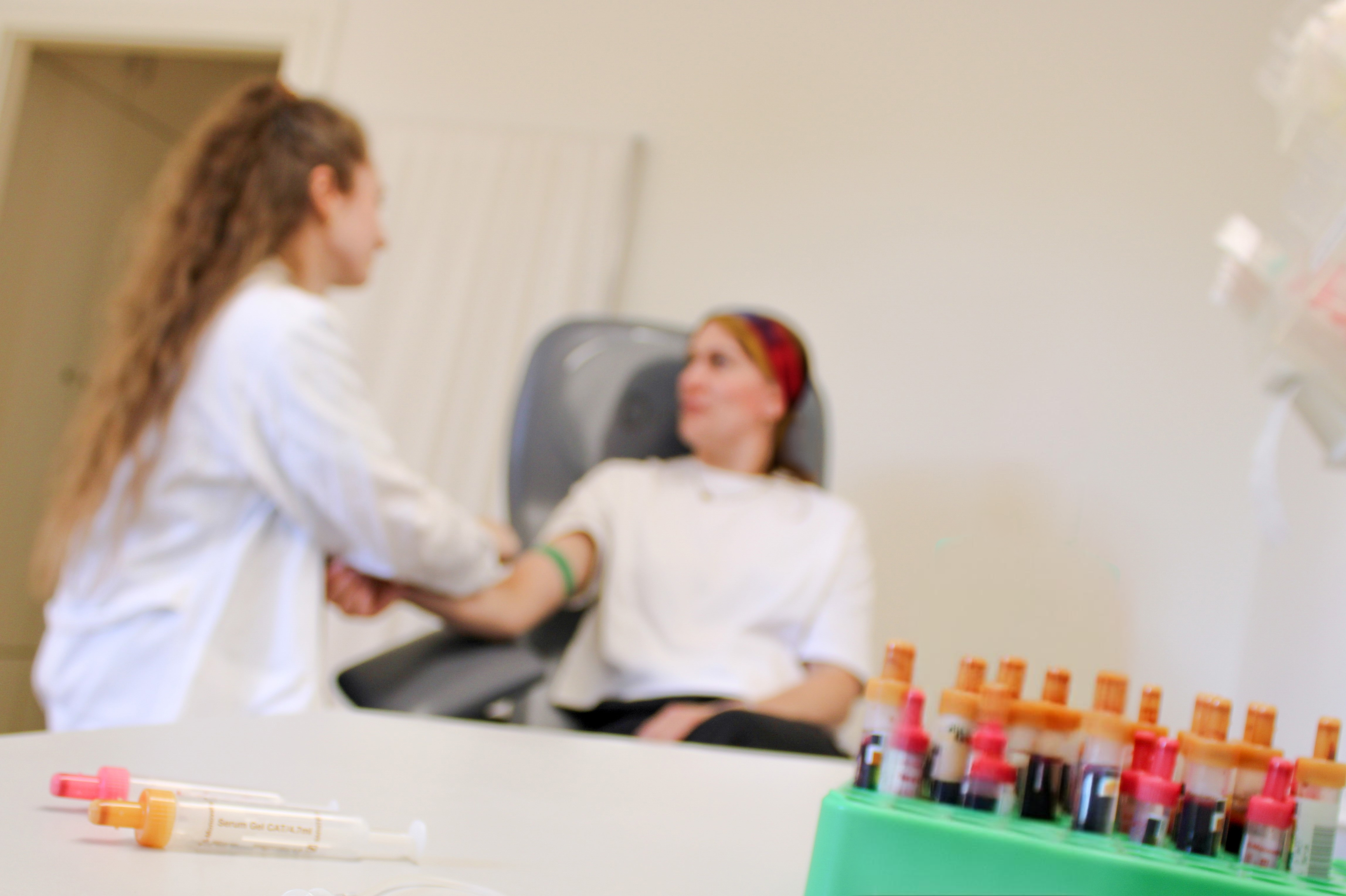 A woman in a white coat prepares to take blood samples from a woman sitting in a treatment chair. In the foreground are full collection tubes in a holder.