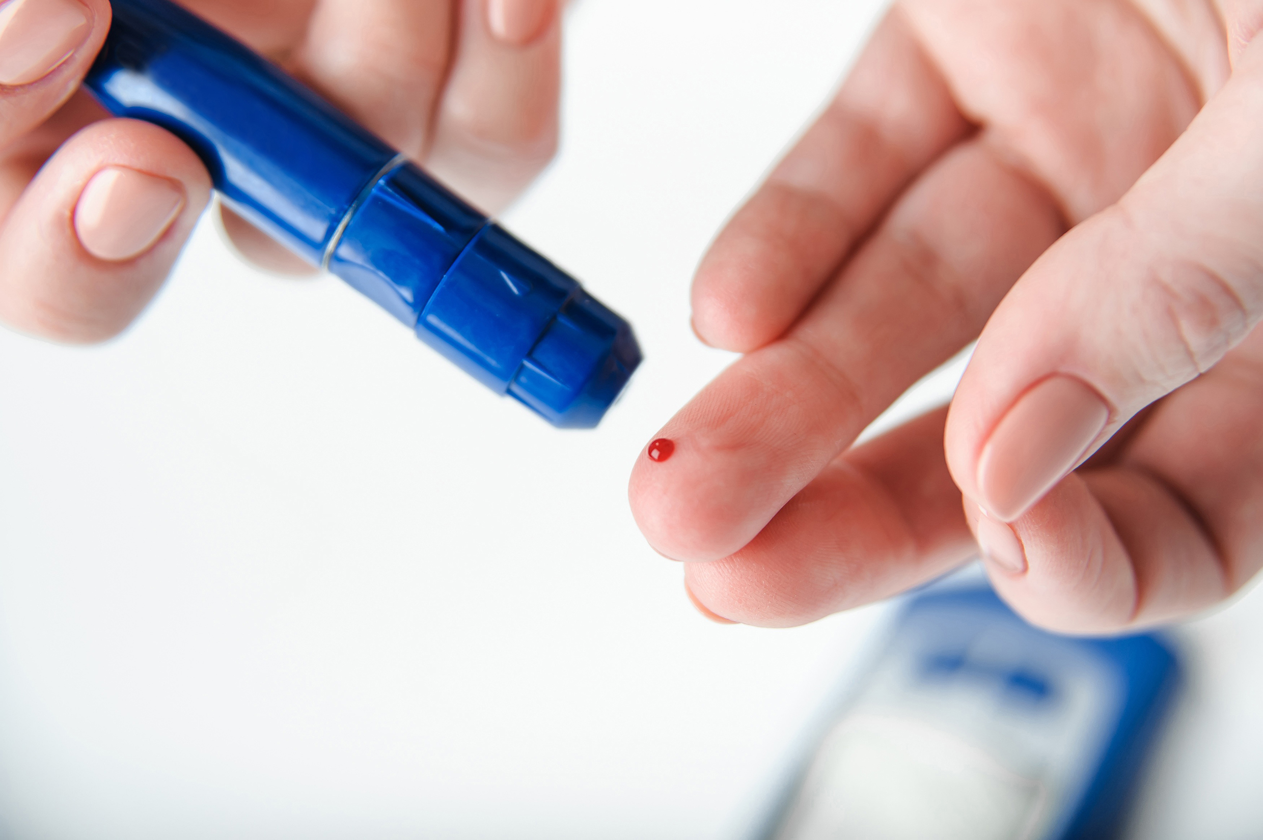 One hand holds a lancing device for measuring blood glucose, the other hand shows a red drop of blood at the puncture site on the ring finger. 