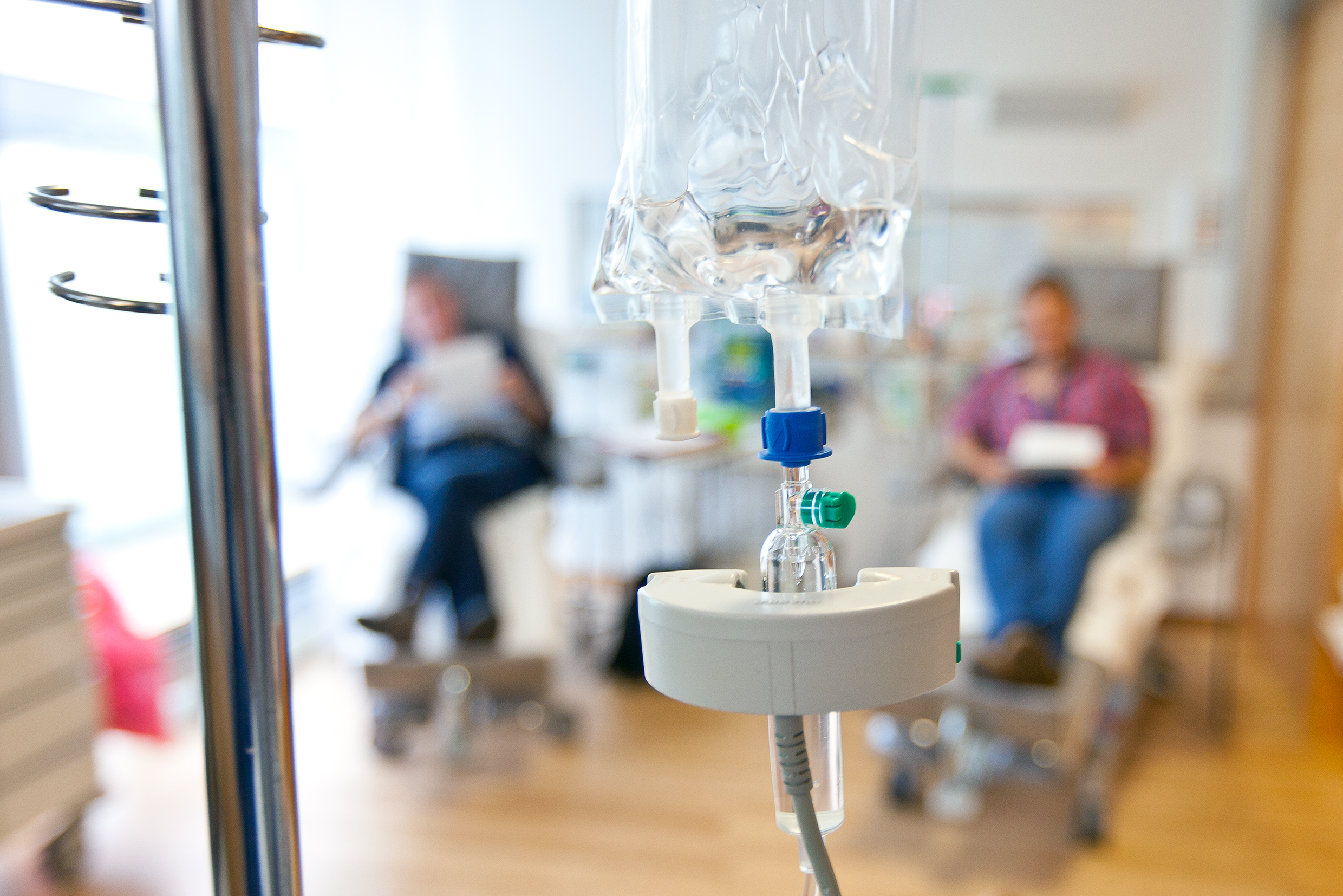 An infusion bag containing a chemotherapy drug hangs in an infusion stand. Two people are sitting in the background.