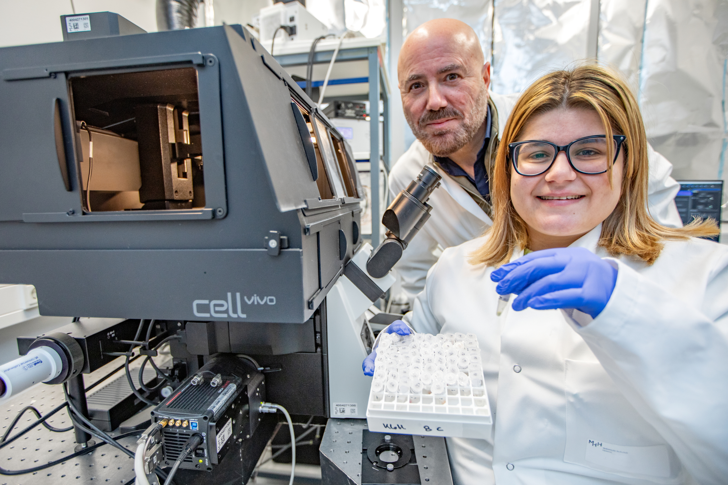 ): A man and a woman are standing next to a special fluorescence microscope and showing a holder with small reaction vessels.