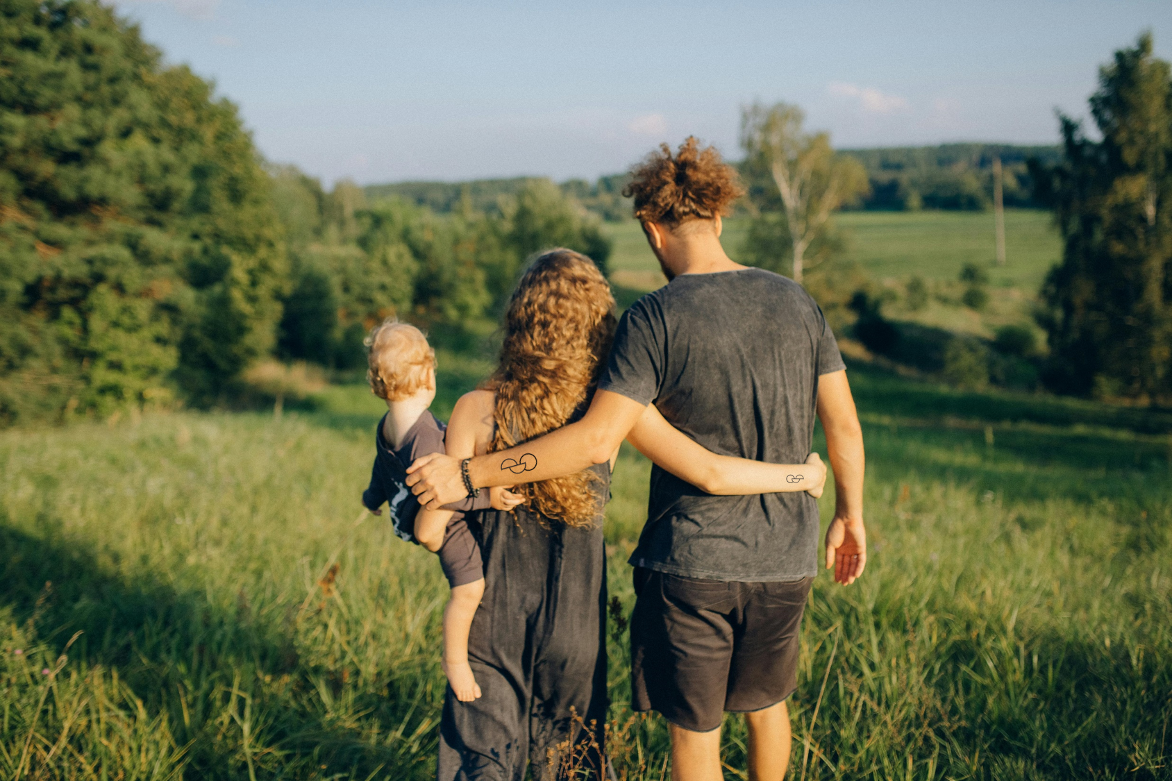 A woman and a man are holding each other, their backs to the camera. The woman is holding an infant. Both woman and man have the organ donation tattoo.