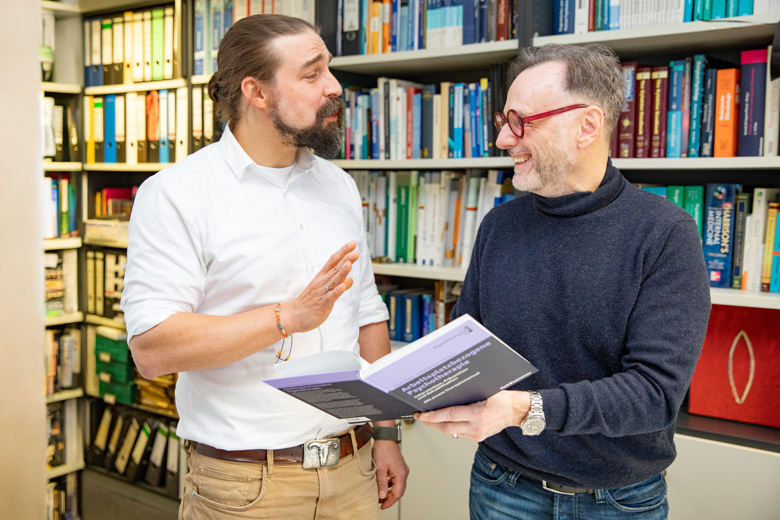 Two men stand in front of a bookshelf with a book in their hands