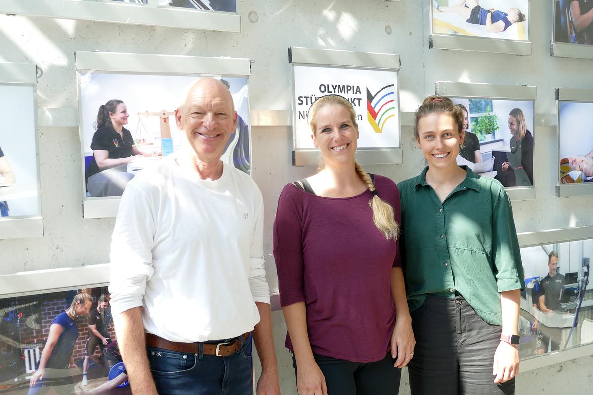 A man and two women are standing in front of a wall of pictures with the words "Olympia Stützpunkt Niedersachsen" (Olympic Training Centre of Lower Saxony).